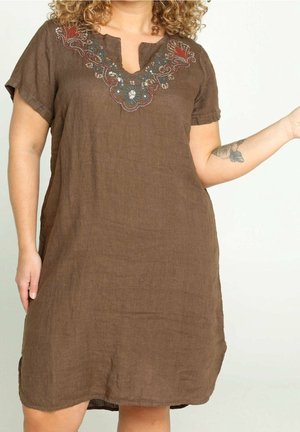 Kvinde iført en kortærmet brun kjole med broderi ved halsudskæringen og en blomstertatovering på underarmen, mod en ensfarvet baggrund.