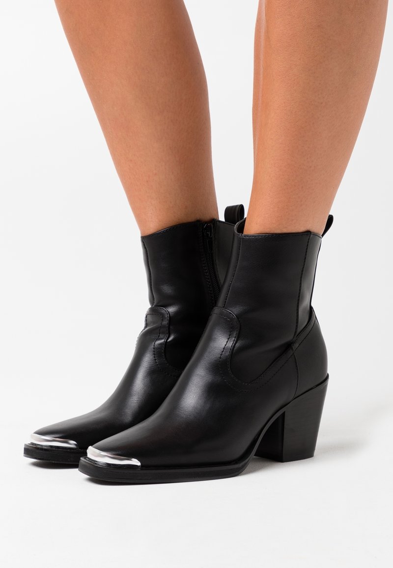 Botines de cuero negro hasta el tobillo con punta afilada, detalles plateados en la puntera, cierre de cremallera y tacón grueso. Textura suave y diseño ajustado.