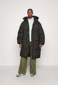 Zwarte pufferjas met een capuchon, met een lange ontwerp, glanzende afwerking, grote gequilte secties en drukknopen. Gecombineerd met groene broek en sneakers.