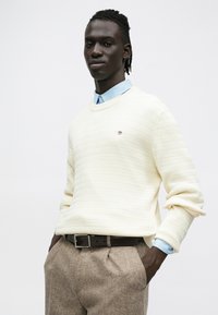 Neulottu kermanvärinen neulepusero, jossa on tekstuuri-kuvio, pyöreä kaula-aukko, yhdistetty vaaleansiniseen kauluspaitaan ja ruskeisiin ruutuhousuihin, joissa on tumma vyö.