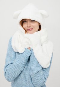Biała polarowa chusta z kapturem z uszami niedźwiedzia, o miękkiej fakturze i zapinanym wzorze. Nosi się ją z jasnoniebieskim swetrem robionym na drutach.