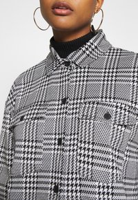 Chemise à motif pied-de-poule noir et blanc en tissu texturé, dotée d'un col, d'une fermeture à boutons et d'une poche poitrine.