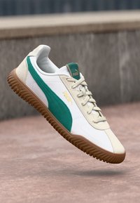 Puma tennis valge ja rohelise disainiga, tekstuurse pruuni tallaga, rippumas neutraalses taustas.