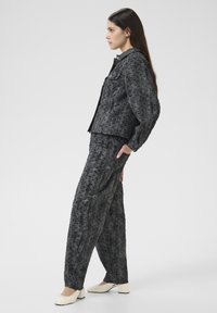 Femme portant une veste texturée gris foncé et un pantalon large assorti, chaussée de chaussures à talon bas beiges, se tenant de profil avec une main dans la poche.