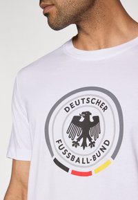 Fehér pamut póló fekete sas logóval, "Deutscher Fussball-Bund" felirattal, valamint szürke, fekete, piros és sárga kör alakú díszítésekkel.