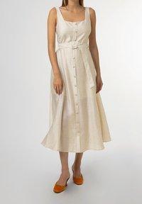 Krémové lněné šaty se čtvercovým výstřihem, zapínáním na knoflíky vpředu a sladěným páskem. Splývavá sukně s mírně do áčka rozšířeným střihem. Kombinováno s oranžovými botami.