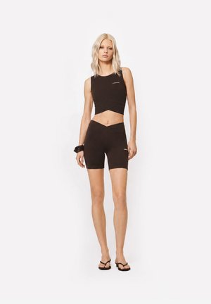 Kobieta o blond włosach, ubrana w czarny bezrękawnik typu crop top i dopasowane czarne krótkie spodenki rowerowe, stojąca na białym tle.