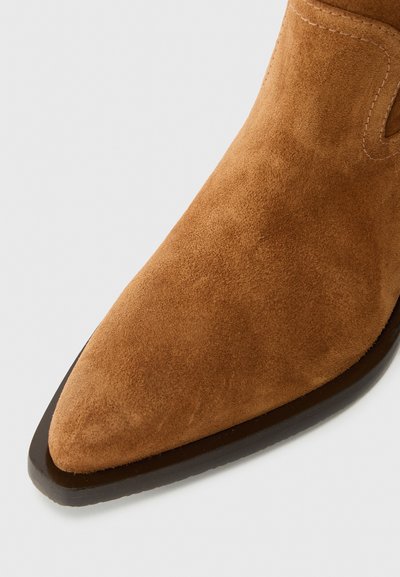 Boots scurte din suede bej, cu vârful ascuțit și talpă subțire maro. Au detalii de cusături pe partea superioară pentru un plus de stil.