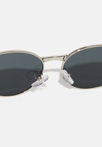 Ochelari de soare ovale cu lentile gri închise, cadru metalic argintiu și suporturi pentru nas din silicon transparent. Design simplu, fără modele sau accente.
