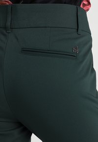 Mörkgröna byxor med en slät textur, en plan bakficka och en liten logotypdetalj nära fickan.
