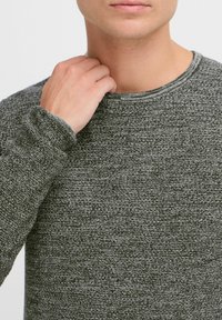 Pulover din tricot gri cu textură, cu guler rotund și mâneci lungi, având o vedere de aproape care evidențiază detaliile materialului și designul manșetelor.