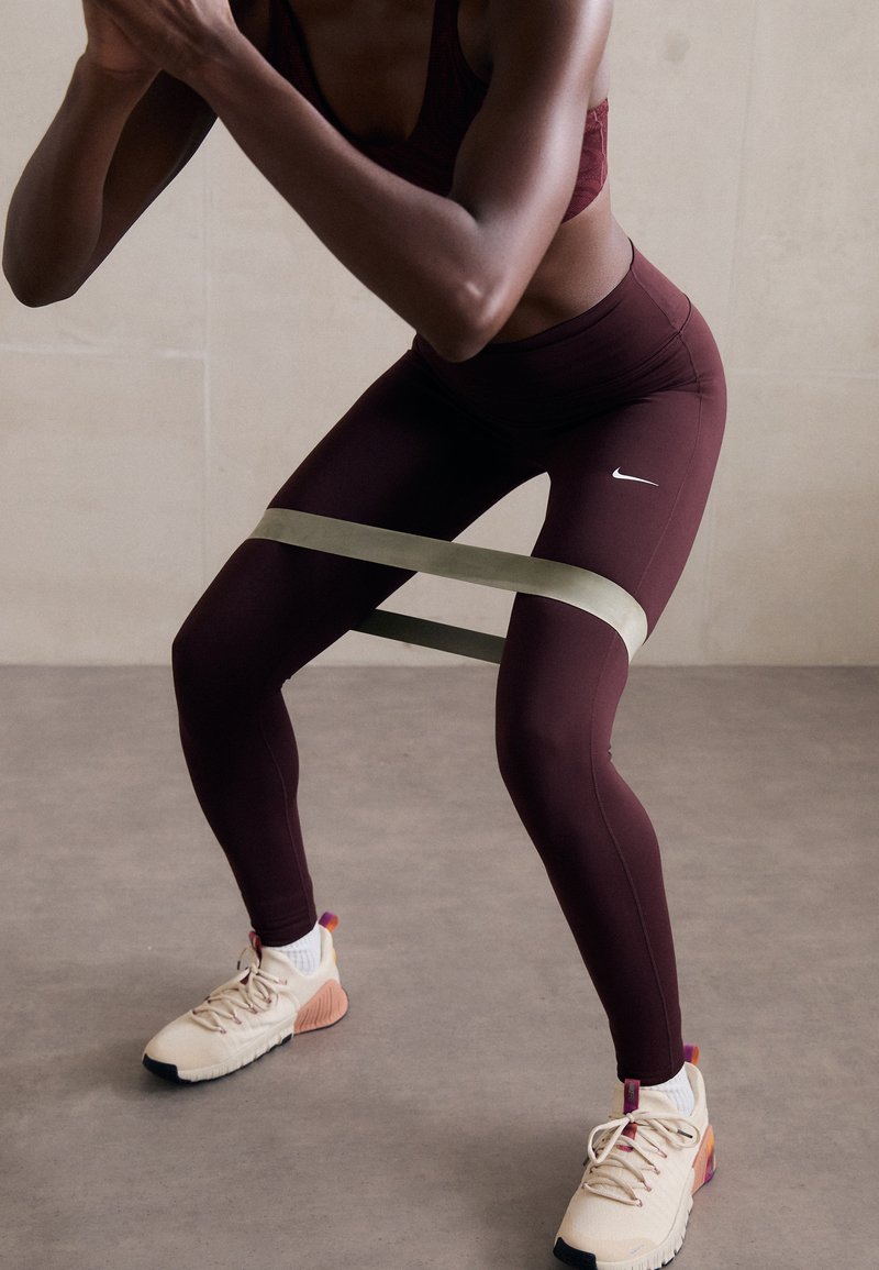 Burgunderfargede leggings med Nike-logo, stylet med et grønt motstandsbånd rundt lårene, kombinert med beige treningssko.