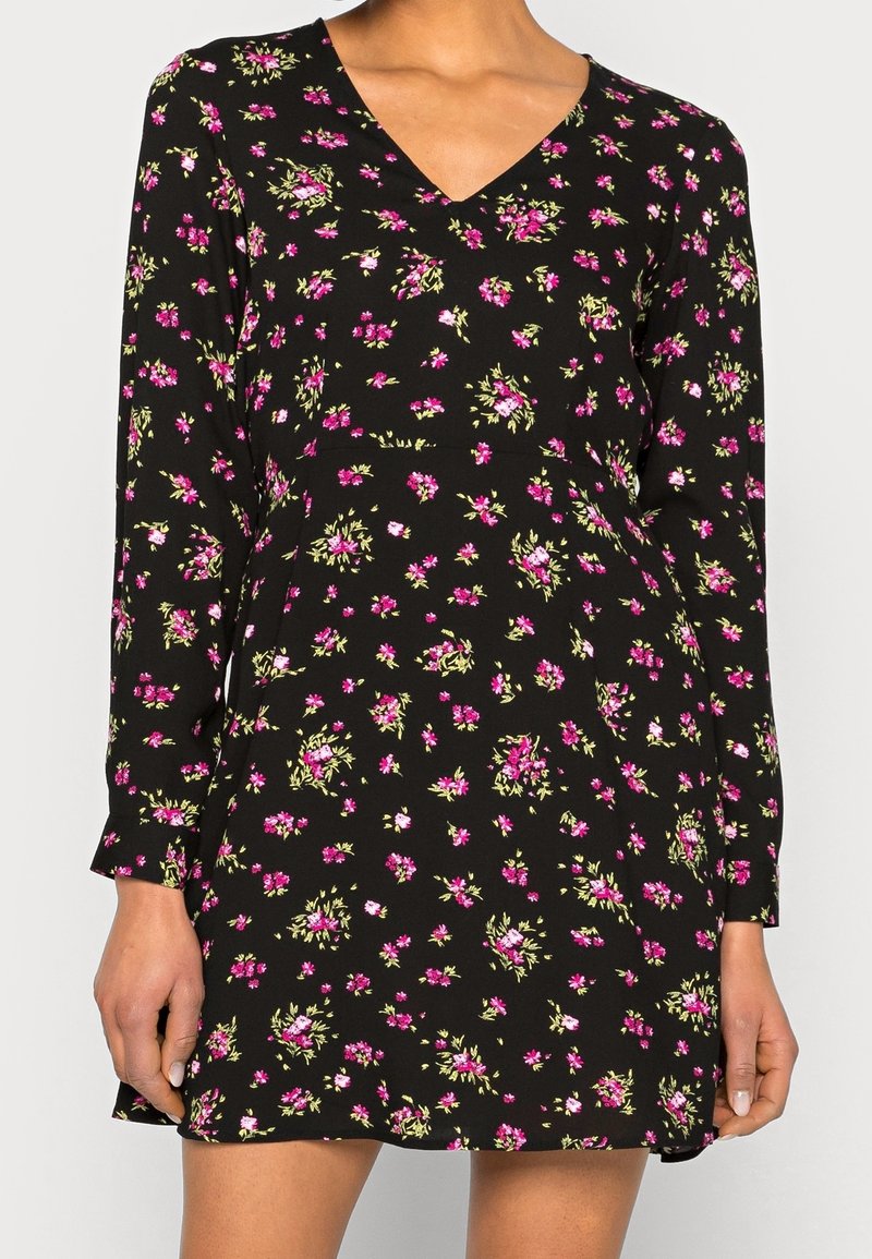 Robe noire à manches longues avec petit motif floral rose et vert et col en V, portée par une femme montrée des épaules jusqu'à mi-cuisse.