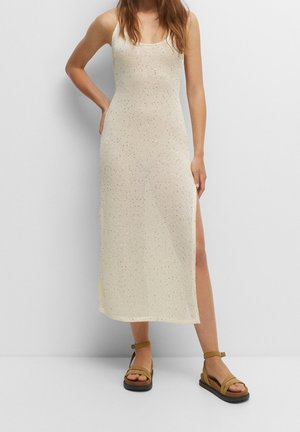 Woman wearing a beige sleeveless knit midi dress with side slit and tan platform sandals against a plain light background.