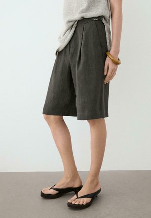 Kvinde iført mørkegrå knælange plisserede shorts, beige striktop, sorte sandaler med hæl og et chunky brunt armbånd på håndleddet.