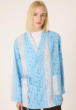 Mladá žena s rovnými blond vlasmi nosí svetlomodrú vzorovanú zavinovaciu bundu previazanú v páse cez biely top.