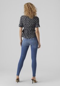 Top nero floreale con maniche corte, design a peplo; abbinato a jeans aderenti blu e sandali neri con tacco alto. Texture morbide, look casual.