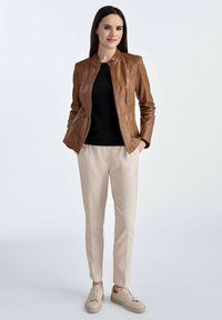 Brązowa skórzana kurtka o dopasowanym kroju, czarna bluzka, beżowe spodnie i jasne sneakersy. Gładka tekstura i kieszenie na zamek.