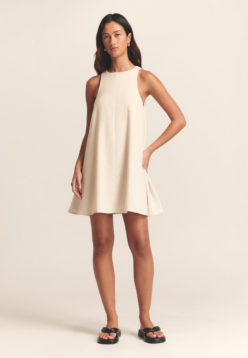 Beige ärmelloses Kleid aus leichtem Stoff mit hohem Ausschnitt, ausgestelltem Saum und seitlichen Schlitzen. Kombiniert mit schwarzen Sandalen.