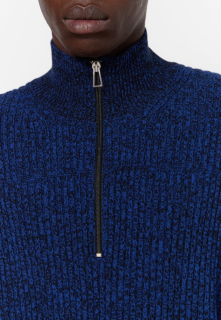 Nahaufnahme einer Person, die einen blau-schwarzen, gerippten Strickpullover trägt, dessen silberner Reißverschluss bis zur Halshöhe geöffnet ist.