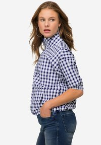 Camicia a quadri blu e bianca con colletto alto e maniche corte, realizzata in tessuto leggero. Indossata con jeans in denim scuro.