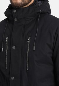 Chaqueta negra con capucha y un acabado texturizado, que presenta bolsillos con cremallera plateada, botones a presión y cordones ajustables en la capucha.