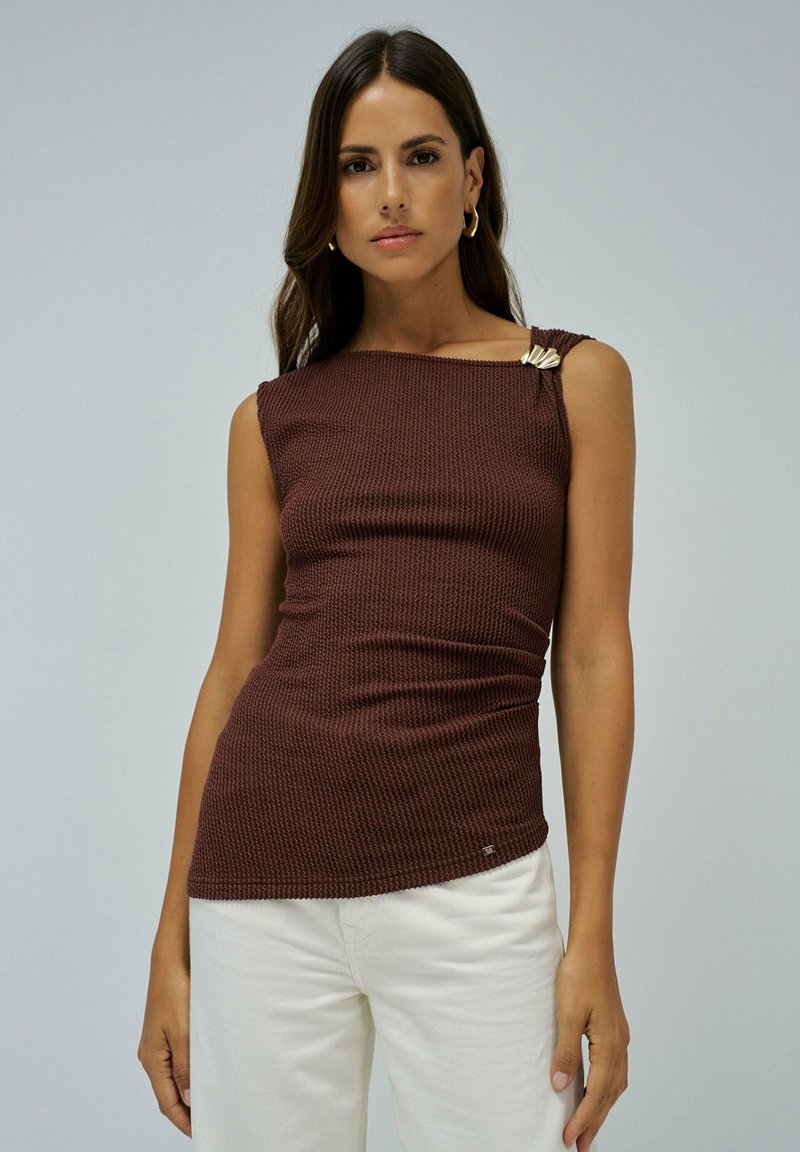 Débardeur en tricot côtelé marron avec un décolleté carré et un accent doré à l'épaule. Associé à un pantalon blanc pour le contraste.