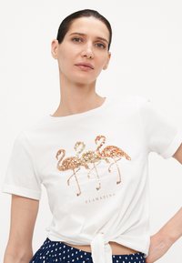 T-shirt blanc en coton avec trois flamants roses en sequins dorés et rose doré, avec un ourlet noué et un col rond.