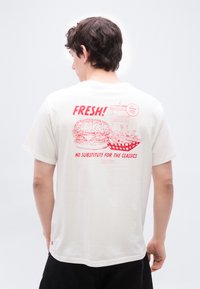 Νεαρός με κοντά σκούρα μαλλιά φορώντας λευκό T-shirt που δείχνει κόκκινο γραφικό με μπέργκερ, πατάτες και ποτό με κείμενο για κλασική φρεσκάδα στην πλάτη.