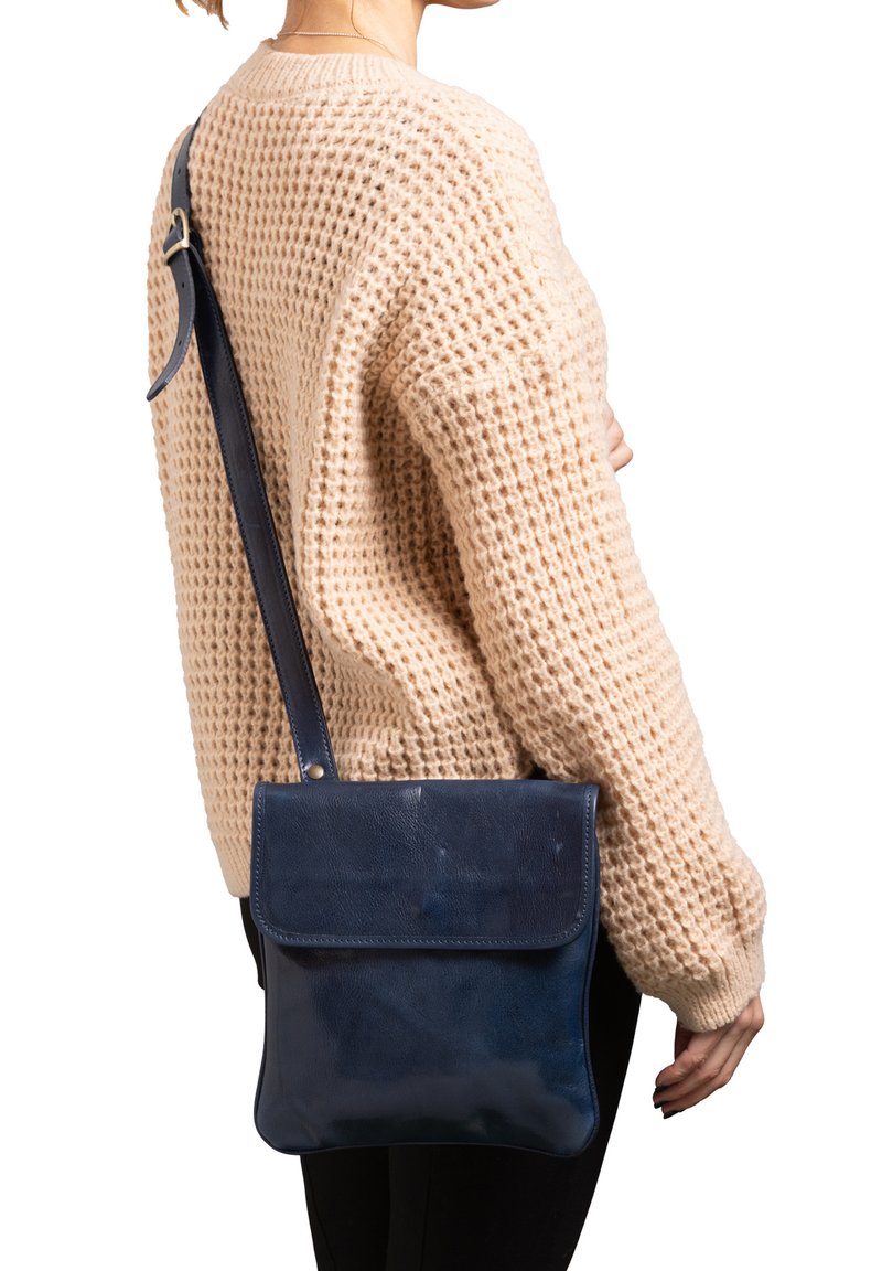 Borsa a tracolla in pelle blu navy con chiusura a pattina, texture liscia e tracolla regolabile, indossata su un pullover in maglia beige.