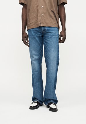 Homme portant un jean bleu ample, une chemise marron à manches courtes avec boutons, et des chaussures habillées noires et blanches, se tenant devant un fond uni.