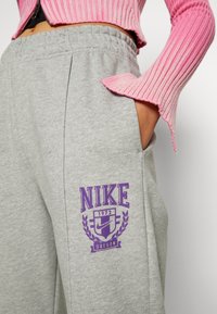 Grå mjukisbyxor med en avslappnad passform, med en lila Nike-logotyp och emblem på vänster ficka. Tillverkad av mjukt bomullsmaterial.