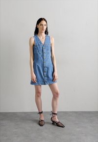 Denim haljina bez rukava s v-izrezom, prednjim zatvaranjem na gumbi i opuštenim krojem. Nošena s crnim potpeticama s remenom oko članka od mrežaste tkanine.
