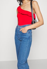 Bodysuit rojo de un solo hombro con detalles de volantes, combinado con jeans de denim azul de tiro alto. La modelo sostiene un pequeño bolso negro.