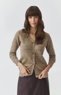 Woman with long brown hair wearing a buttoned beige cardigan and dark skirt, adjusting the bottom button on a plain light background.