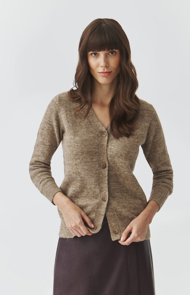 Woman with long brown hair wearing a buttoned beige cardigan and dark skirt, adjusting the bottom button on a plain light background.