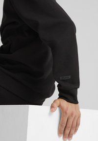 Sudadera negra hecha de material suave con un diseño ajustado. Presenta un pequeño logo gris de Puma cerca del puño y una tela texturizada.