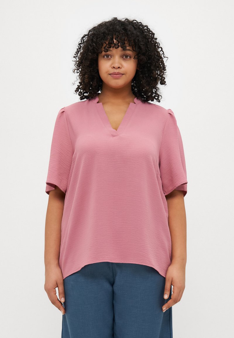 Pink short-sleeve blouse with a V-neck and subtle texture, paired with loose blue trousers. Hemline is curved at the bottom.