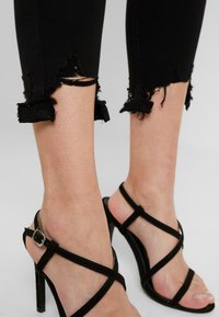 Černé poškozené džíny s třepícími se lemy, doplněné černými sandály na vysokém podpatku s pásky, které mají sponu a hladký povrch.