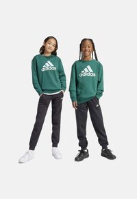 Felpa verde in cotone con logo Adidas bianco, abbinata a pantaloni della tuta neri. I modelli indossano sneaker bianche e nere, mostrano uno stile sportivo e casual.