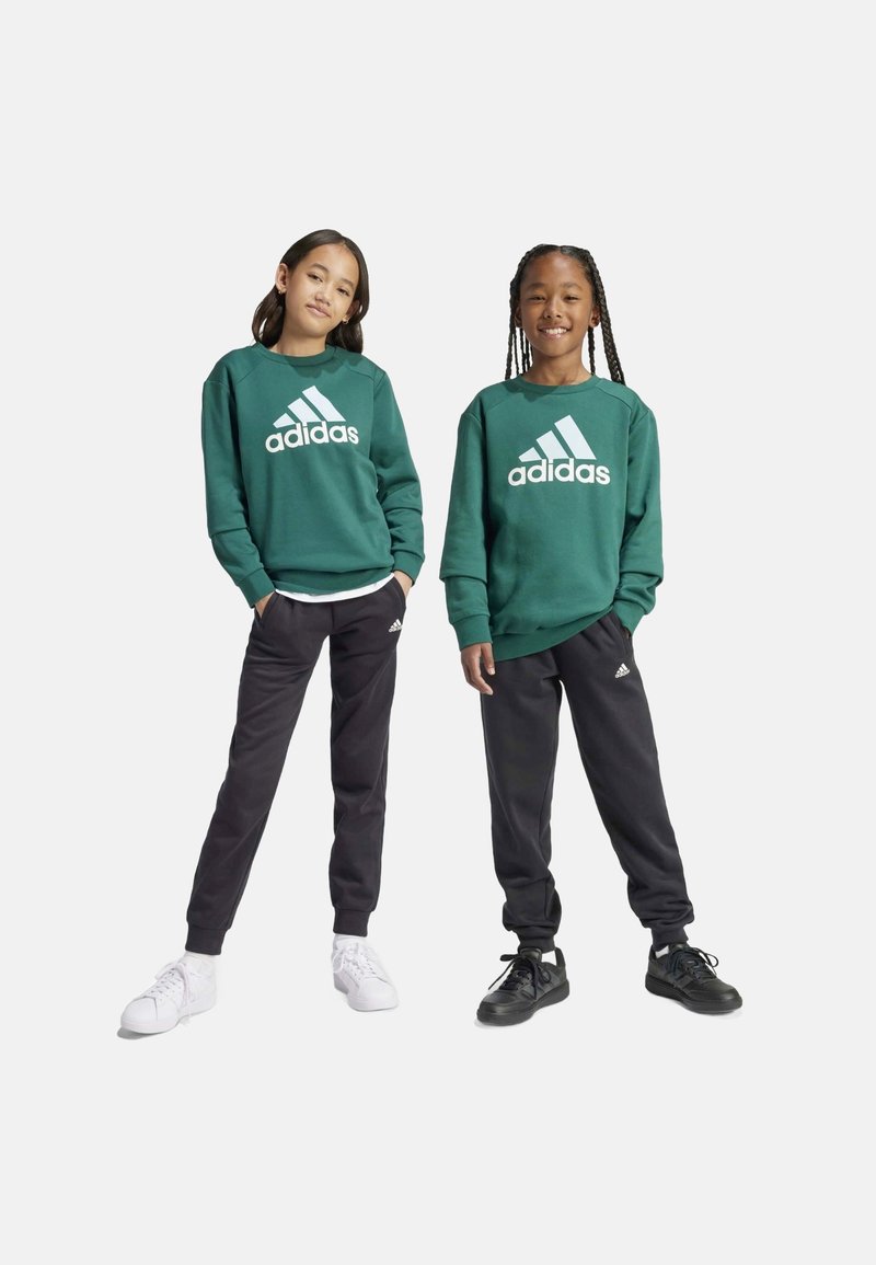 Felpa verde in cotone con logo Adidas bianco, abbinata a pantaloni della tuta neri. I modelli indossano sneaker bianche e nere, mostrano uno stile sportivo e casual.