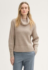 Beżowy sweter z dzianiny z kołnierzem, prążkowanym wzorem i splotem warkoczowym na dole. Połączony z jasnoszarymi dopasowanymi spodniami.