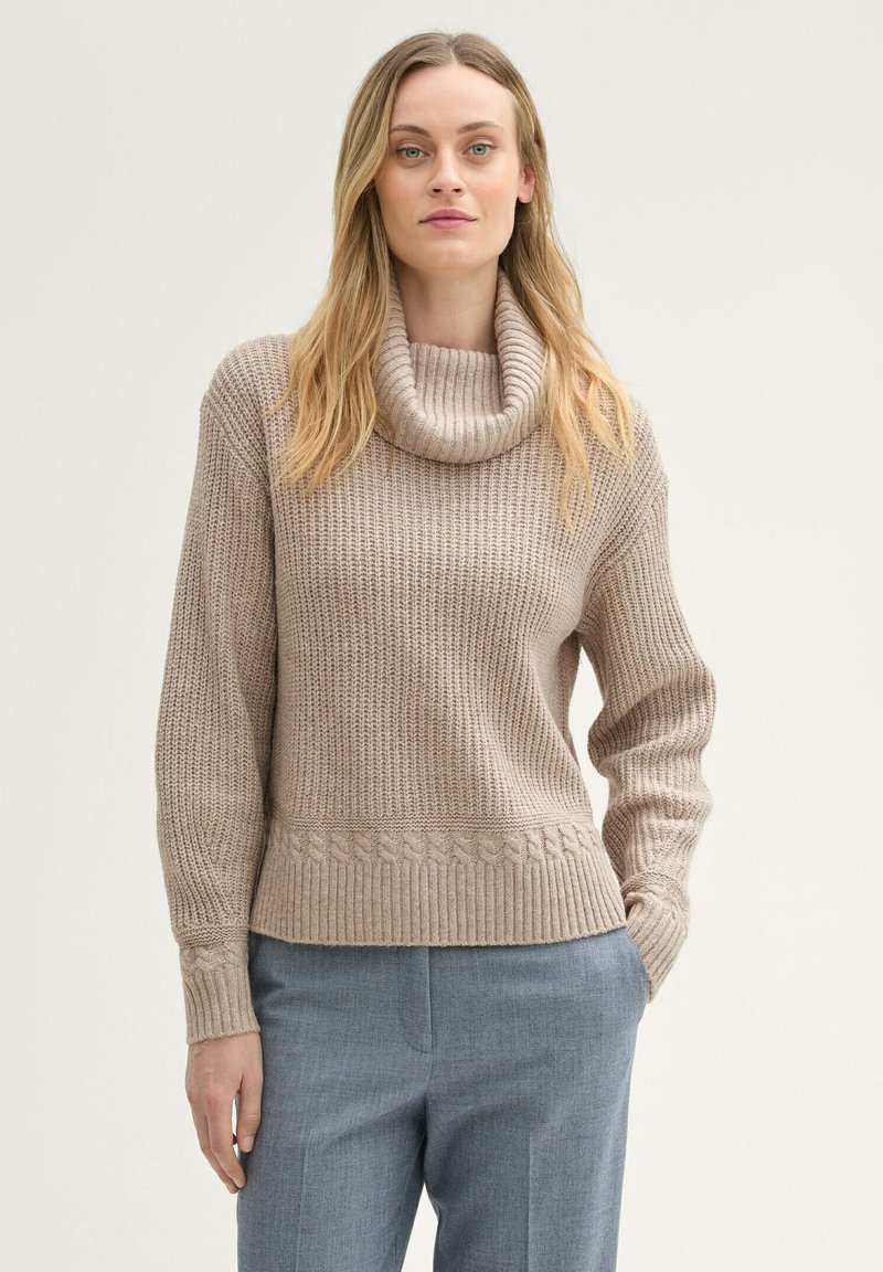 Beżowy sweter z dzianiny z kołnierzem, prążkowanym wzorem i splotem warkoczowym na dole. Połączony z jasnoszarymi dopasowanymi spodniami.