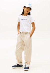 Λευκό μπλουζάκι Tommy Hilfiger με μπλε λογότυπο, μπεζ παντελόνι φαρδιάς γραμμής και μπλε ναυτικό αθλητικά με λευκές σόλες. Λευκό καπέλο με μπλε διακοσμητική λεπτομέρεια.