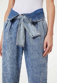 Pantalones de mezclilla con una cinturilla ancha y atada. Color azul claro, dobladillo despuntado y diseño con pliegues, mostrando un acabado de tela texturada.