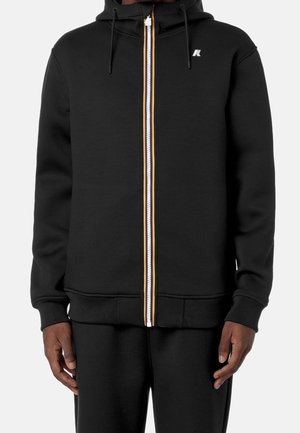 Czarna bluza z kapturem na zamek błyskawiczny z białym i pomarańczowym paskiem na zamku, sznurki przy kapturze oraz małe białe logo na piersi, noszona przez osobę.