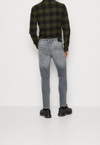 Vaqueros grises de corte ajustado con un lavado desgastado, diseño de cinco bolsillos y parche trasero con el logo. Combinados con una camisa de franela a cuadros en negro y oliva.