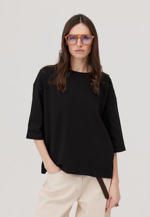 Kobieta z długimi brązowymi włosami, nosząca przesadnie duże pomarańczowe okulary z niebieskim odcieniem, czarną luźną koszulę i beżowe spodnie, stojąca na jasnym tle.