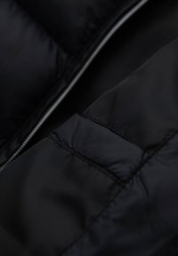 Tissu en duvet noir avec une texture lisse et brillante. Présente une fermeture éclair visible et un quilting, offrant isolation et structure.