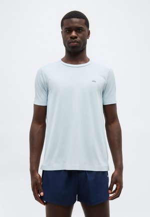 Jauns vīrietis, tērpies gaiši zilā sporta krekla ar īsām piedurknēm un tumši zilās šortos, stāv pret vienkāršu fonu.